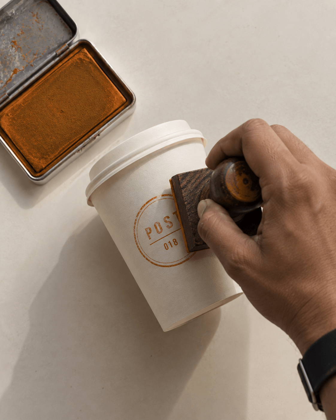 A hand pressing a wooden rubber stamp onto a Post coffee cup beside an open ink pad.