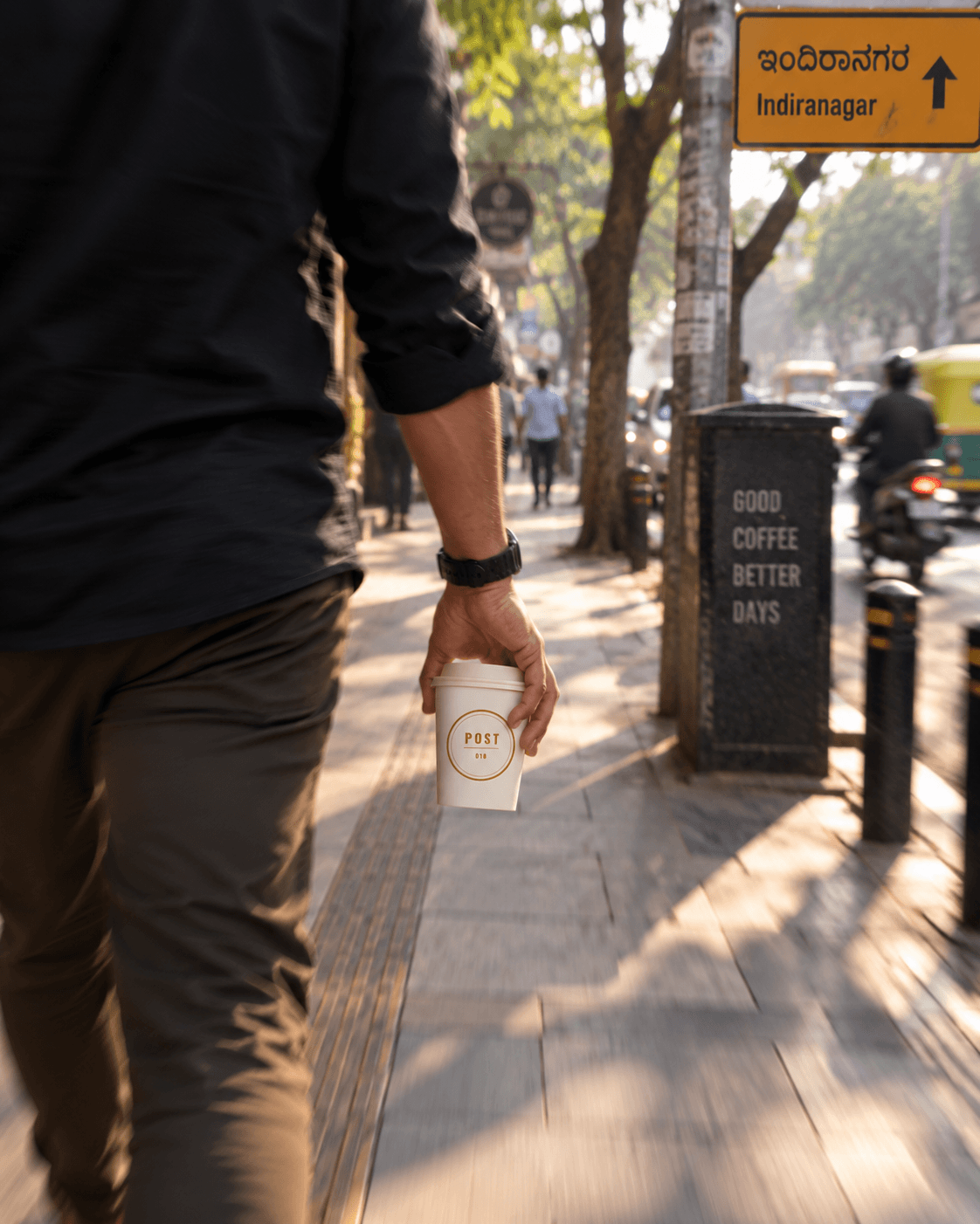 A man walking down an Indiranagar footpath carrying a Post coffee cup.